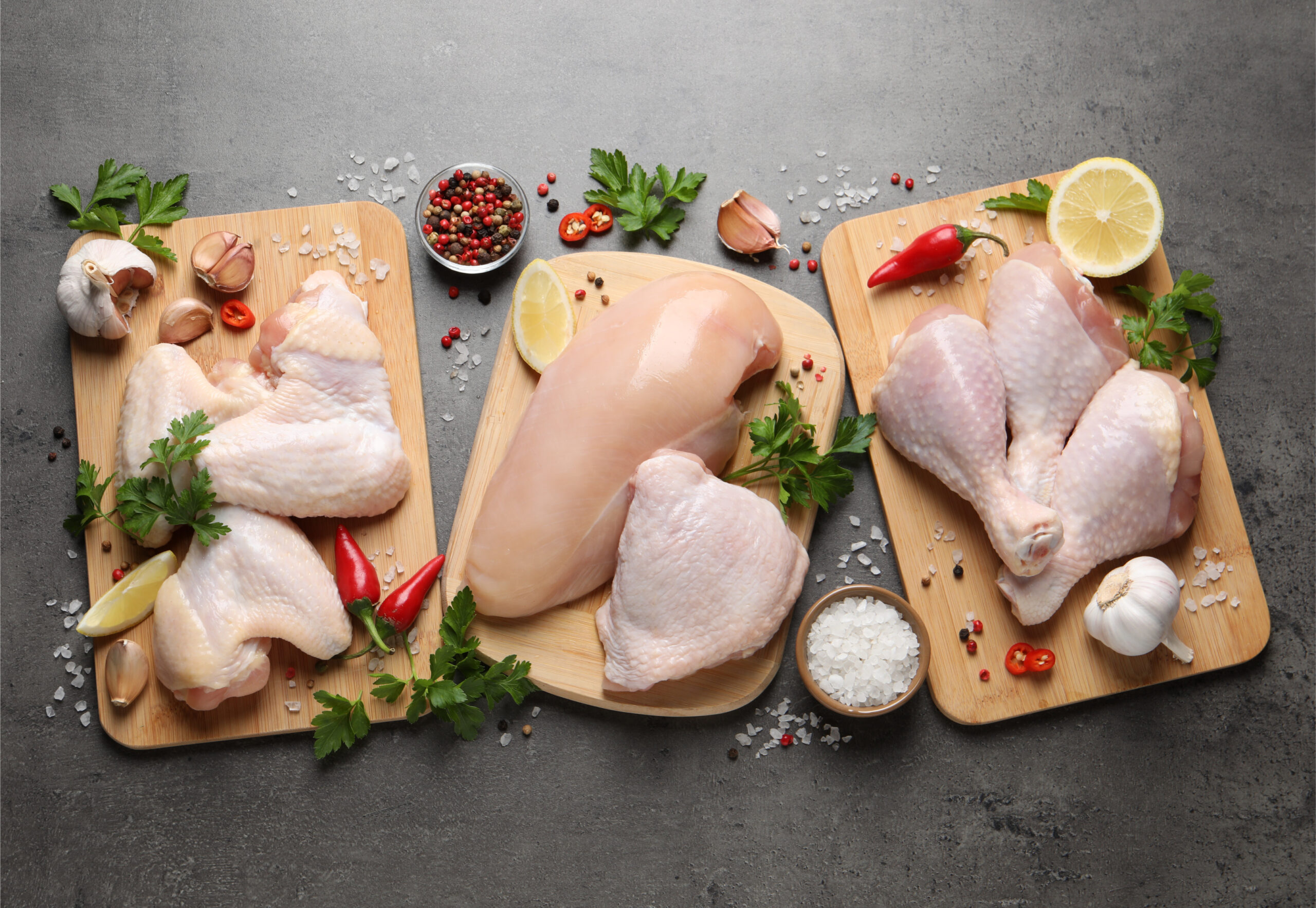 Raw chicken pieces including breasts, thighs, and drumsticks arranged on wooden cutting boards, garnished with fresh parsley, garlic, chili peppers, and lemon slices, set against a dark gray background.