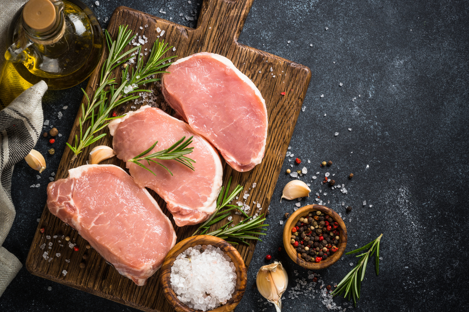 Fresh pork chops on a wooden cutting board, surrounded by garlic, rosemary, and a mix of colorful peppercorns, with olive oil and sea salt in the background, ideal for a delicious meal preparation.
