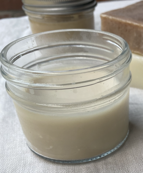 Close-up of a glass jar filled with creamy homemade moisturizer, set against a textured background with a bar of soap in the background. Ideal for skincare enthusiasts and DIY beauty product creators.