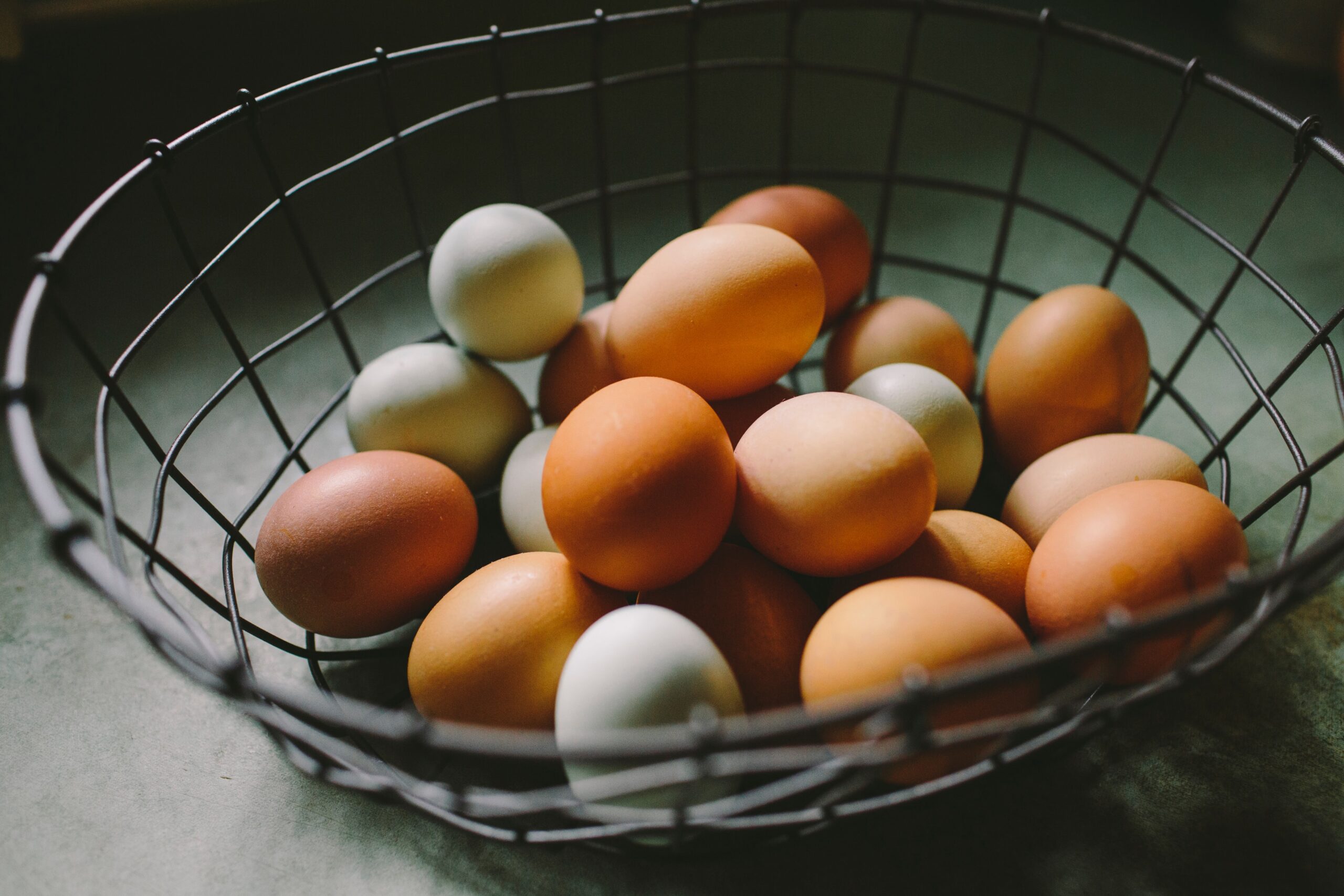 A wire basket filled with a variety of eggs, including brown and pale green, resting on a textured surface, emphasizing natural colors and farm-fresh produce.